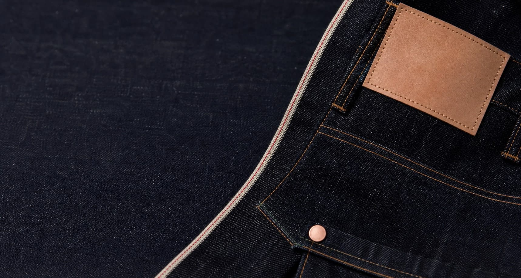 Macro de denim selvedge escuro com linha vermelha, rebite de cobre e patch de couro sem marca.